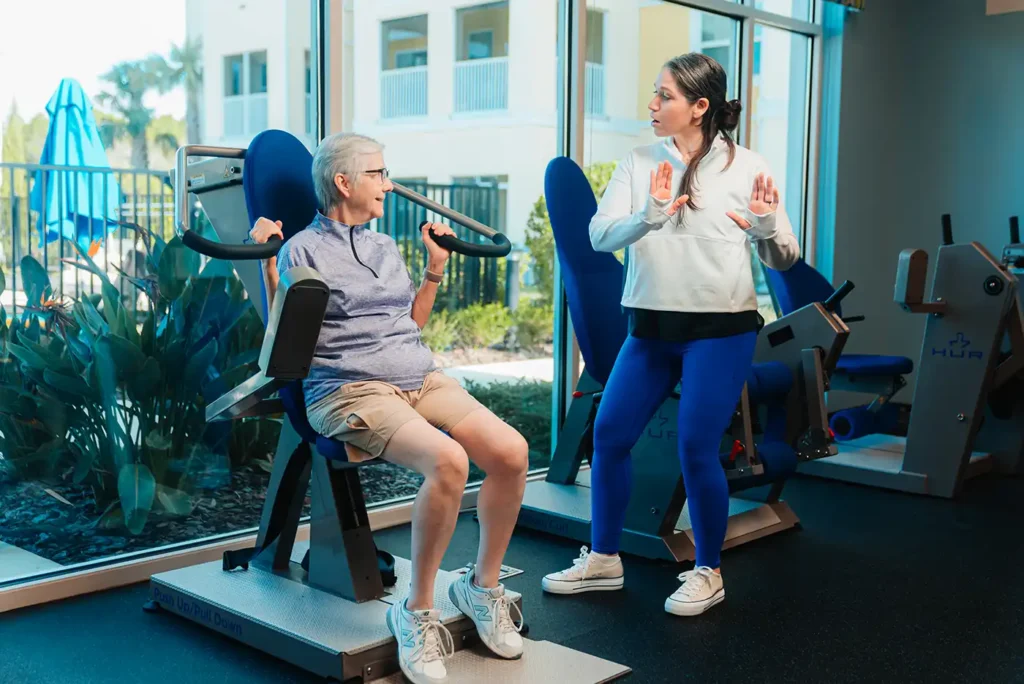 A senior woman prepares to use a piece of gym equipment while her trainer mimics correct posture